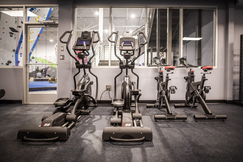 Photo of Inner Peaks Fitness Facility showing stationary bikes and ellipticals with a view of climbing walls in the background. Click to visit our Fitness Facility page.